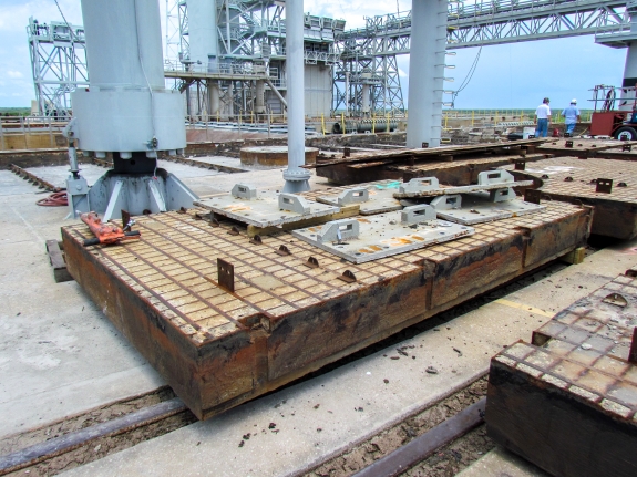 Large-format photograph: Pad Deck, Launch Complex 39-B, east side of Flame Trench. Removed Crawlerway Grid Panels lie stacked on cribbing, with Side Flame Deflectors Hold-down Lugs and Base Plate segments stacked on top of them. Just beyond them, you can see MLP Mount Mechanism 3 (left), one of the MLP Rainwater Downspouts (center), and a Sound Suppression Water Header (right), which feeds SSW Water up into the underside of the MLP. Farther distant, across the Flame Trench on its west side, you can see Pad Infrastructure for the Artemis Program, the Space Shuttle Program, and original Apollo Program, all mixed together. Photo credit: Withheld by request.